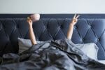 person in bed under covers with mug making victory sign